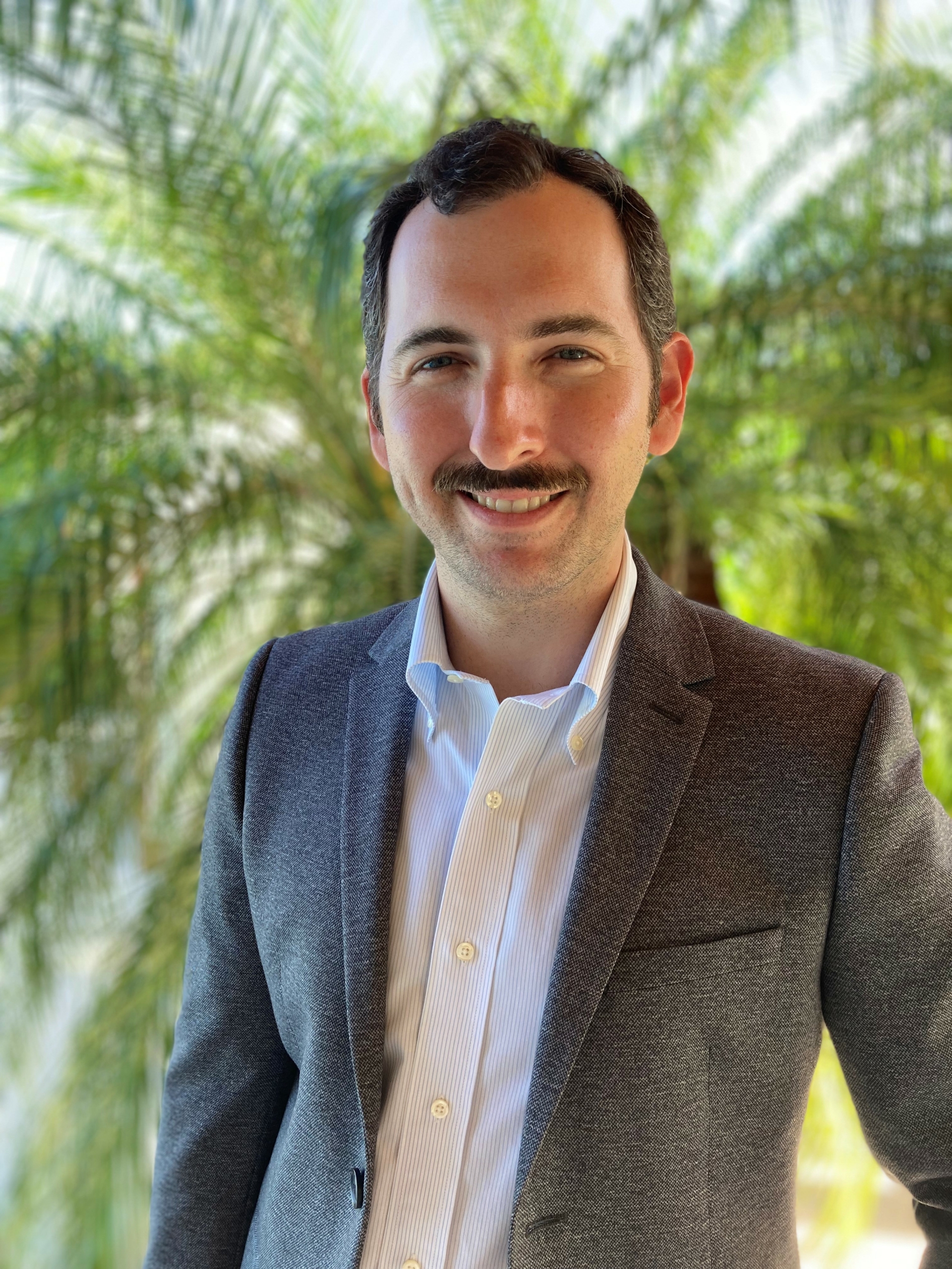 A man with a mustache, dressed in a gray blazer and white shirt, stands in front of palm trees with a slight smile on his face.