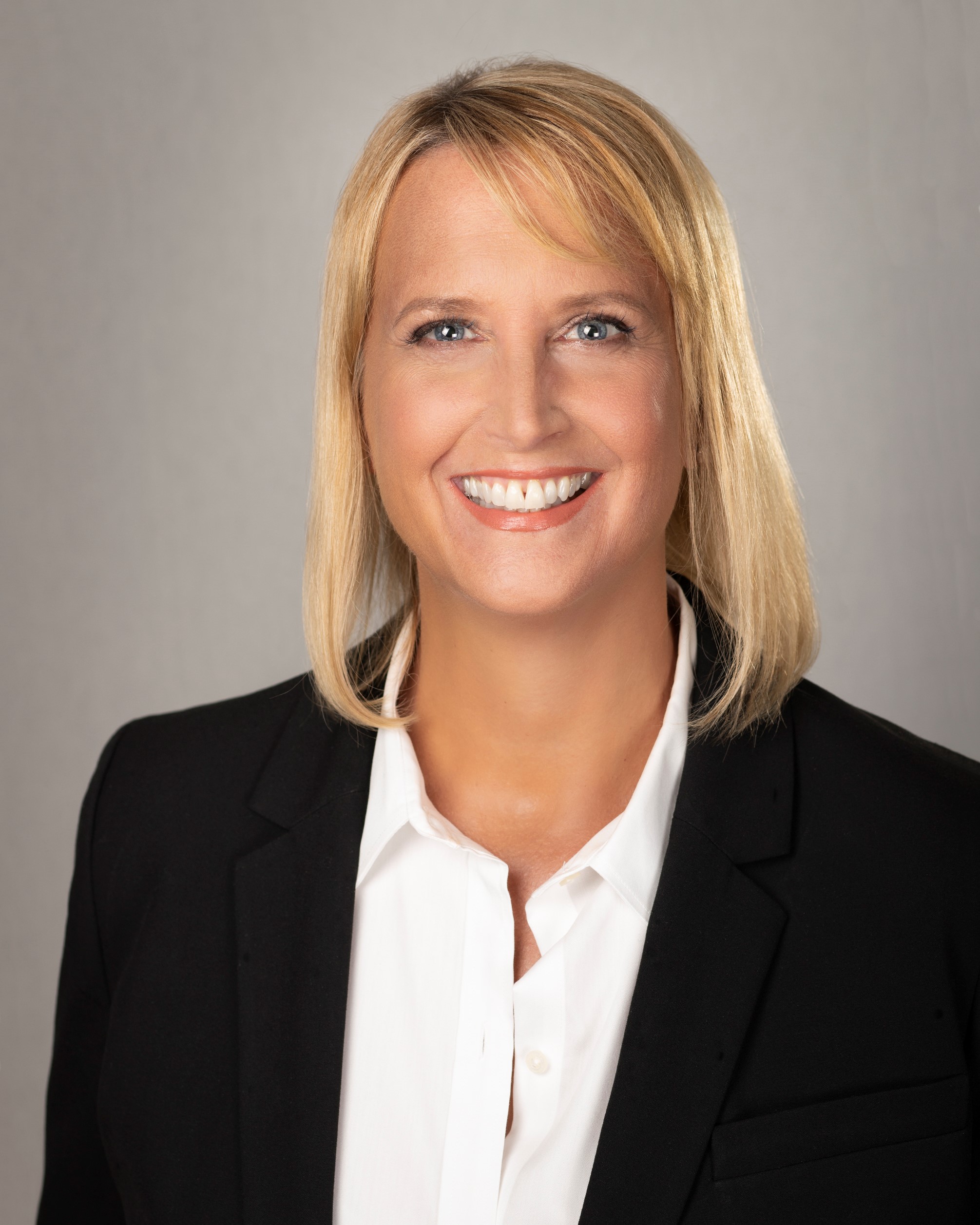 A person with shoulder-length blonde hair smiles, wearing a black blazer and white shirt, against a gray background.