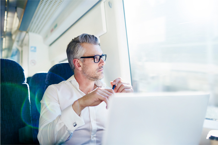 A man with gray hair and glasses sits on a train, looking out the window while holding a pen with an open laptop in front of him.