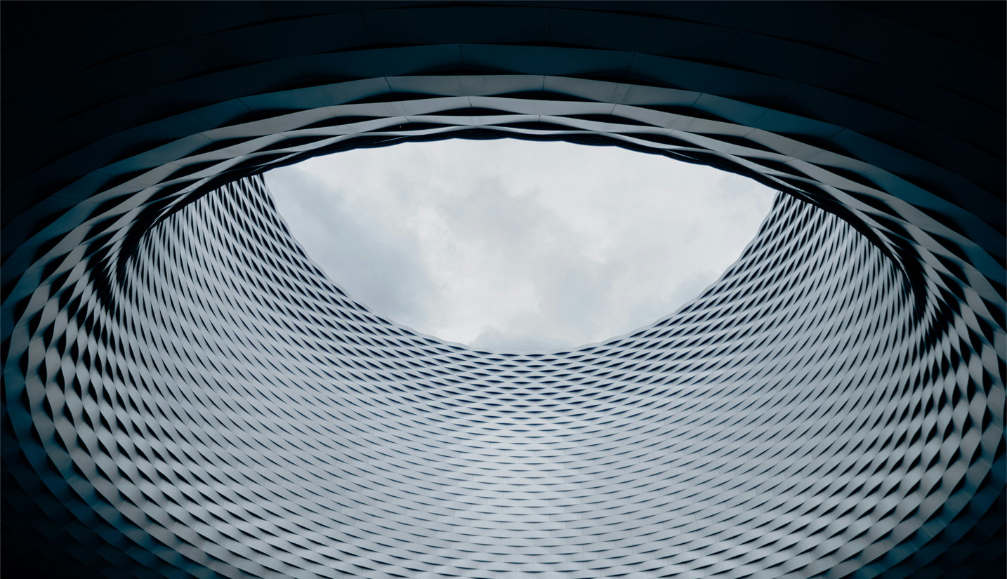 Upward view of a modern architectural structure with a circular opening at the top, allowing a glimpse of the sky. The design features a pattern of interlocking metallic panels.