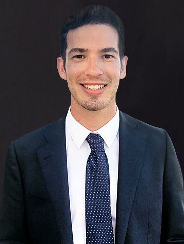 Man in a dark suit and polka dot tie smiling against a dark background.