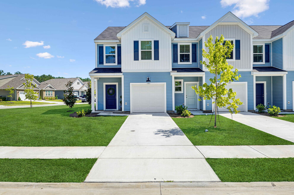 A row of modern townhouses with blue and white exteriors, small front yards, driveways, and young trees in a suburban neighborhood on a sunny day.