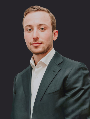 A man with short light brown hair, wearing a dark suit jacket and a white dress shirt, poses against a plain dark background.