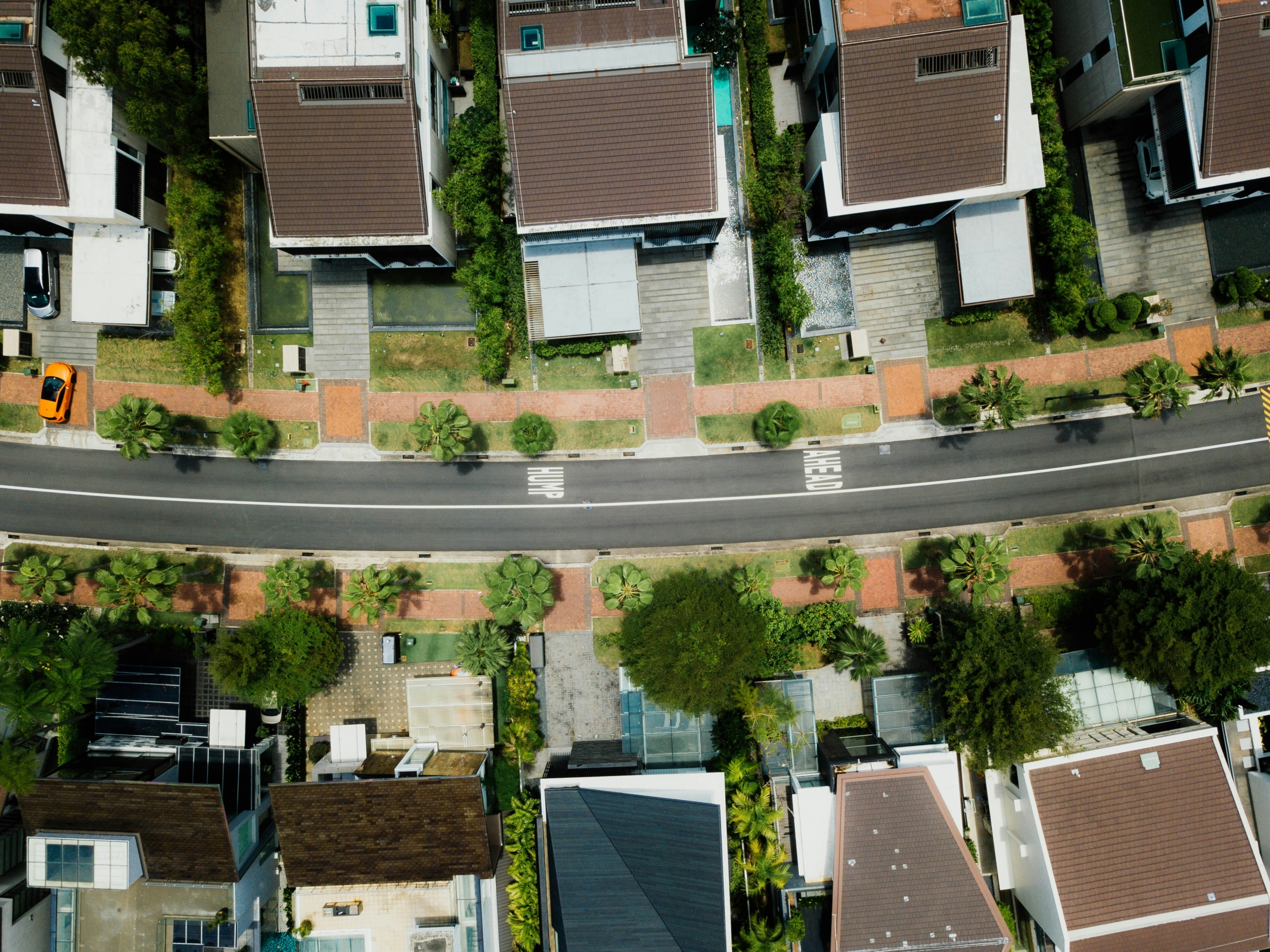 Aerial view of a suburban neighborhood with houses lined along a curved street. A single orange car is parked on the road. Green lawns and trees are visible in the area.